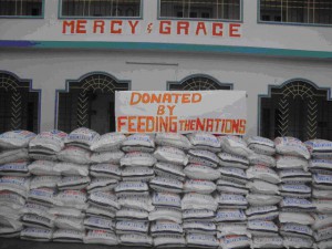 in fornt pf Mercy and Grace Building, a sign above dozens of bags of food says, " Donated by Feeding The Nations"